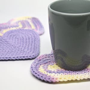 May include: A gray ceramic mug resting on a purple and yellow crocheted heart-shaped coaster. Two additional heart-shaped coasters are visible in the background.