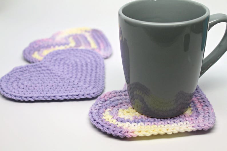 May include: A gray ceramic mug resting on a purple and yellow crocheted heart-shaped coaster. Two additional heart-shaped coasters are visible in the background.