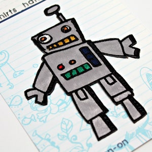 Pode incluir: Um remendo de ferro de passar roupa de robô cinza com contornos pretos, uma boca amarela, olhos laranja e um painel de luz vermelho e verde. O remendo está sobre um fundo branco com linhas azuis e rabiscos.