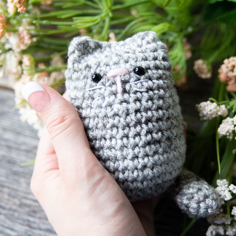 May include: A handmade gray crochet cat with black eyes, a pink nose, and white whiskers. The cat is held in a hand, with a blurred background of greenery and flowers. The cat has a small tail.