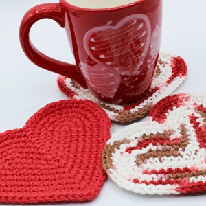 May include: Three crocheted heart-shaped coasters in red, white, and brown. A red mug with a white heart design sits on top of one of the coasters.