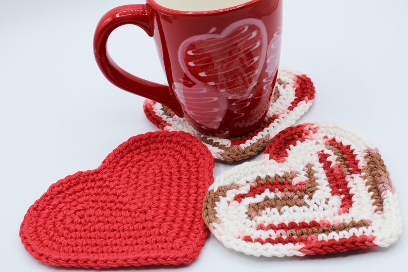 May include: Three crocheted heart-shaped coasters in red, white, and brown. A red mug with a white heart design sits on top of one of the coasters.