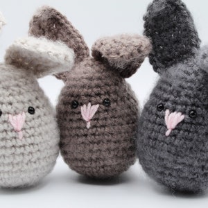 May include: Three crocheted bunny plushies in different colors: light gray, brown, and gray. Each bunny has a pink nose and black eyes.