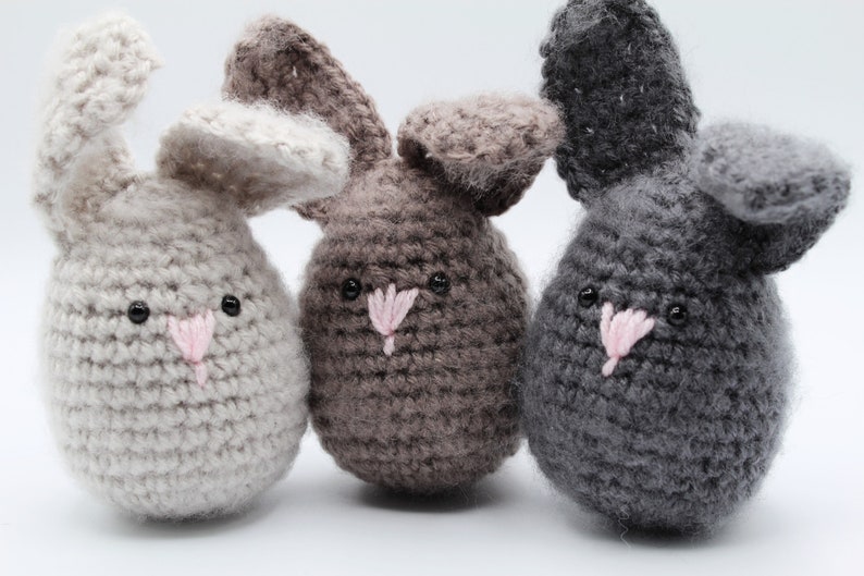 May include: Three crocheted bunny plushies in different colors: light gray, brown, and gray. Each bunny has a pink nose and black eyes.