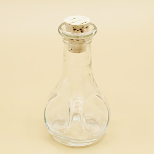 Op de afbeelding: Een heldere glazen fles met een kurken stop. De fles heeft een unieke, bolle vorm met een getextureerd oppervlak.