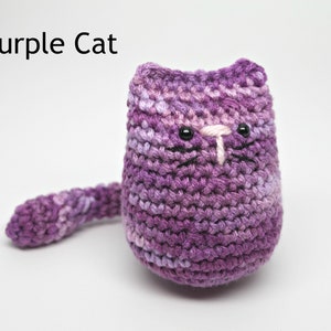 May include: A handmade, crocheted cat in shades of purple. The cat has black eyes, a white nose, and black whiskers. The text "Purple Cat" is above the cat. The cat has a long tail.