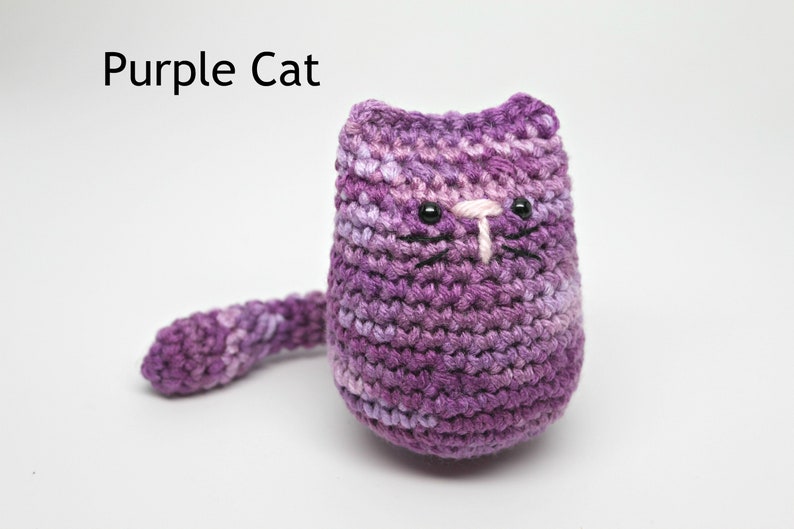 May include: A handmade, crocheted cat in shades of purple. The cat has black eyes, a white nose, and black whiskers. The text "Purple Cat" is above the cat. The cat has a long tail.