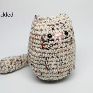 May include: A speckled, handmade crochet cat doll. The cat is beige with flecks of color, featuring black eyes, whiskers, and a pink nose. The word "Speckled" is in the upper left corner.