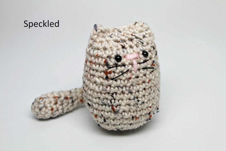 May include: A speckled, handmade crochet cat doll. The cat is beige with flecks of color, featuring black eyes, whiskers, and a pink nose. The word "Speckled" is in the upper left corner.