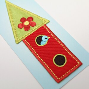 Op de afbeelding: Een rode en groene vilt vogelhuis applique met een blauwe vogel en een rode bloem op het dak. De applique is op een lichtblauwe achtergrond.