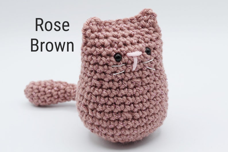 May include: A crocheted cat toy in rose brown with glitter yarn. The cat has black eyes and a pink nose with white whiskers.
