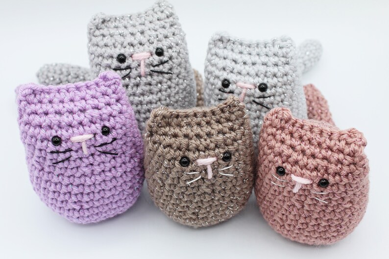 May include: Five crocheted cat toys in different colors, including purple, gray, brown, and pink. Each cat has black eyes, a pink nose, and black whiskers.