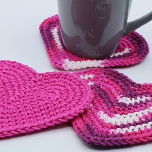 May include: Three crocheted heart-shaped coasters, two in pink and one in shades of pink, purple, and white. A gray ceramic mug sits on top of one of the coasters.