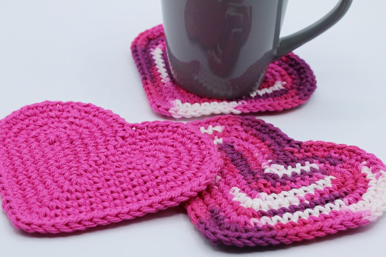 May include: Three crocheted heart-shaped coasters, two in pink and one in shades of pink, purple, and white. A gray ceramic mug sits on top of one of the coasters.