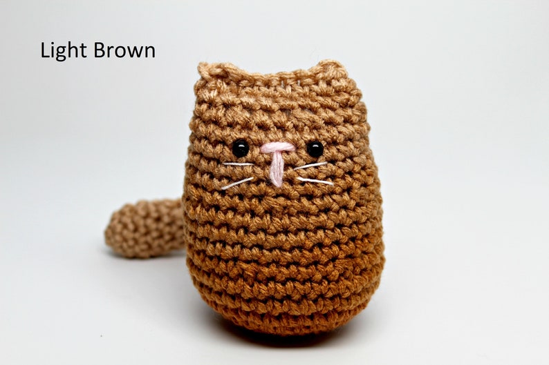May include: A crocheted cat doll in shades of light brown. The handmade cat has black eyes, white whiskers, and a pink nose. The text "Light Brown" is in the upper left corner. The cat has a small tail.