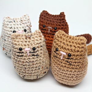 May include: Four handmade crochet cat toys in various colors: cream, brown, tan, and light brown. Each cat has black eyes, pink noses, and black whiskers. The toys are made with textured yarn and have small tails.