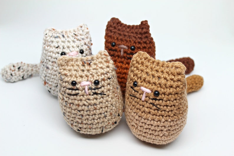 May include: Four handmade crochet cat toys in various colors: cream, brown, tan, and light brown. Each cat has black eyes, pink noses, and black whiskers. The toys are made with textured yarn and have small tails.