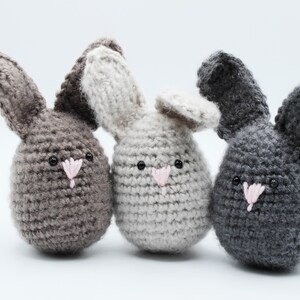 May include: Three crocheted bunny plushies in different colors: gray, light gray, and dark gray. Each bunny has a pink nose and black eyes.