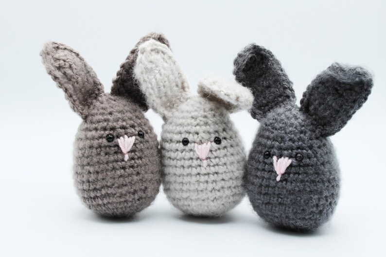 May include: Three crocheted bunny plushies in different colors: gray, light gray, and dark gray. Each bunny has a pink nose and black eyes.