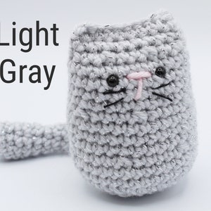 May include: A light gray crocheted cat with black eyes, a pink nose, and black whiskers. The cat has a long tail.