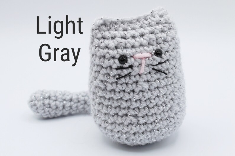 May include: A light gray crocheted cat with black eyes, a pink nose, and black whiskers. The cat has a long tail.