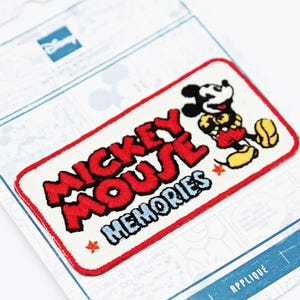 Op de afbeelding: Een rechthoekige patch met Mickey Mouse en de woorden "Mickey Mouse Memories" in het rood. Mickey draagt zijn klassieke outfit. De patch staat op een witte achtergrond met een rode rand en is bevestigd aan een Disney-merkrug.
