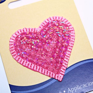 May include: Pink heart-shaped sequin patch with a pink satin border. The patch is on a brown card with blue and white waves and the text "Aplicacion".