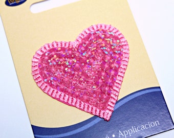 Puede incluir: Parche de lentejuelas en forma de corazón rosa con un borde de satén rosa. El parche está en una tarjeta marrón con ondas azules y blancas y el texto "Aplicacion".