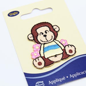 May include: Embroidered monkey patch on a cream card. The monkey has brown fur, a tan face, and a blue and white striped shirt. Pink flowers are on either side of the monkey. The card has the word "Wrights" at the top.