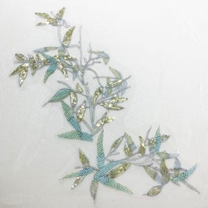 Könnte beinhalten: Gestickte florale Applikation mit einem Astdesign mit Blättern. Die Blätter sind eine Mischung aus goldenen und hellblauen Pailletten und Perlen, mit silbernen Fadenästen. Die Applikation befindet sich auf weißem Hintergrund.