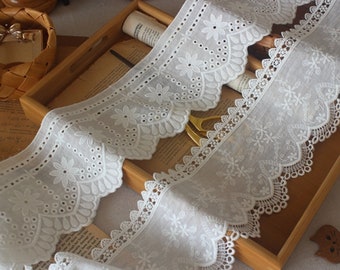 May include: Two white cotton lace trims with scalloped edges and floral embroidery. The trim on the top has larger, more spaced out flowers and holes. The trim on the bottom has smaller, more densely packed flowers and holes.