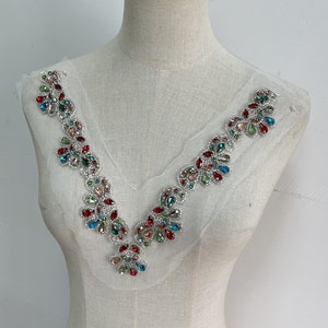 May include: A colourful rhinestone applique with a V-neck design. The applique is made of silver metal and features red, blue, green, and clear rhinestones.
