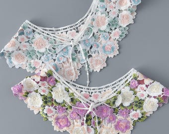 May include: Two white lace collars with floral embroidery. The top collar features light blue and pink flowers, while the bottom collar has purple and pink flowers.