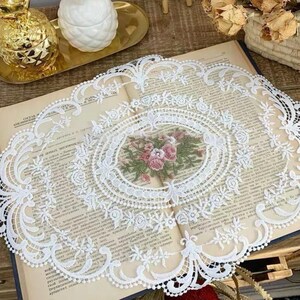 Può includere: Tovaglietta di pizzo bianco con un motivo floreale al centro. La tovaglietta è posizionata sopra un libro dall'aspetto vintage.