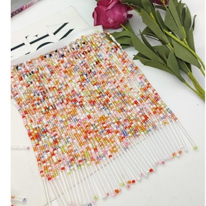 Puede incluir: Una sección de flecos de cuentas con un respaldo blanco. Los flecos están densamente llenos de pequeñas cuentas de colores en tonos de rojo, naranja, amarillo, verde, rosa y blanco. Los hilos transparentes cuelgan, creando un efecto texturizado.