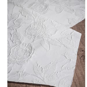 May include: Close-up of white fabric with a raised floral rose pattern. The textured design features detailed roses and leaves, creating a three-dimensional effect. The fabric is set against a wooden surface.