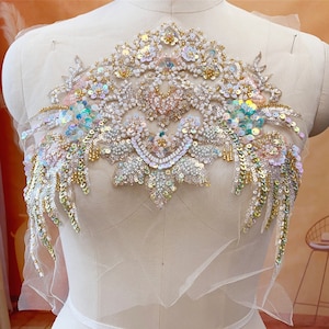 Puede incluir: Aplicación bordada y adornada con cuentas, con un diseño floral. La aplicación presenta lentejuelas, cuentas y perlas iridiscentes en tonos dorados, blancos y pastel. Montada sobre un soporte de tela blanca transparente.