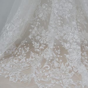 May include: White floral lace fabric with a delicate pattern and a subtle shimmer. The lace is soft and flowing, making it perfect for a variety of projects.