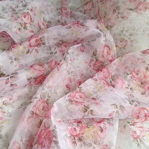 May include: A sheer white fabric adorned with a floral design of pink roses and tiny yellow blossoms. The fabric is arranged in soft folds, creating shadows. The overall effect is romantic and airy, suitable for craft projects.