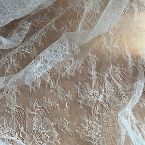 May include: Close-up of white lace fabric with a floral pattern. The delicate lace features detailed designs of flowers and leaves, creating a textured and elegant appearance. The fabric is draped, showing the intricate details of the lace.