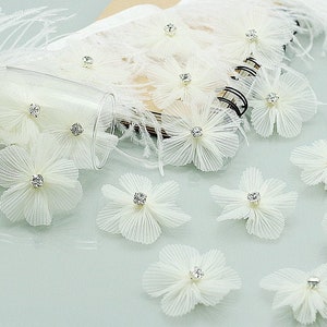 May include: White fabric flowers with a rhinestone center. The flowers are arranged on a white surface with a notebook and feathers in the background.