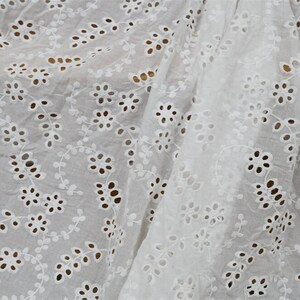 Könnte beinhalten: Weißer Baumwollstoff mit einem zarten Ösenmuster aus kleinen Blüten und Blättern. Der Stoff ist durchscheinend und hat eine weiche, fließende Textur.