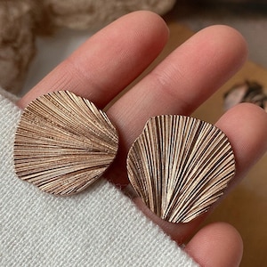 Może przedstawiać: Para złotych kolczyków z wzorem w kształcie wachlarza. Kolczyki są duże i mają fakturowaną powierzchnię.