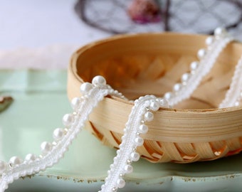 May include: White beaded trim with a delicate lace pattern. The trim is made of small white beads and a thin white lace. The trim is shown on a light blue surface.
