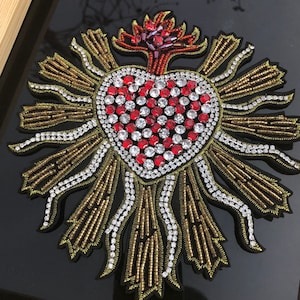 May include: A red heart with a gold and white beaded halo, adorned with red and clear rhinestones. The heart is surrounded by a halo of gold and white beads.