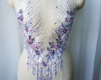 May include: A delicate white lace applique with purple floral embroidery and pearl accents. The applique features a cascading design with dangling pearl strands.
