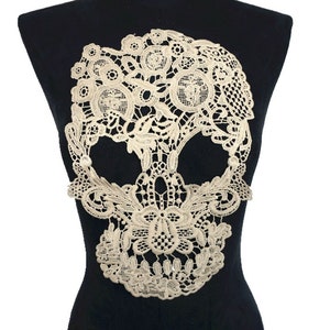 May include: Ivory lace skull applique. The skull design is made of intricate floral and circular patterns. The applique is displayed on a black mannequin, showcasing the detailed openwork of the lace. Ideal for adding a unique touch to clothing or accessories.