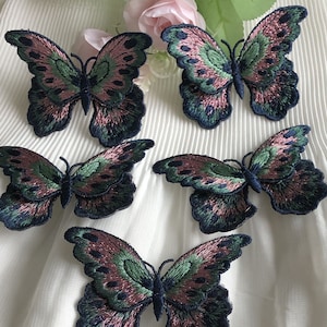 Può includere: Cinque toppe ricamate a forma di farfalla in tonalità di verde, rosa e nero. Le farfalle sono disposte a semicerchio su uno sfondo di tessuto bianco.