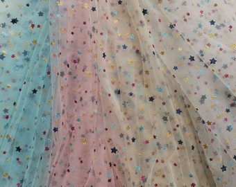 May include: A close-up of a sheer fabric with a star pattern. The fabric is in three colors: light blue, pink, and cream. The stars are gold, blue, and red.