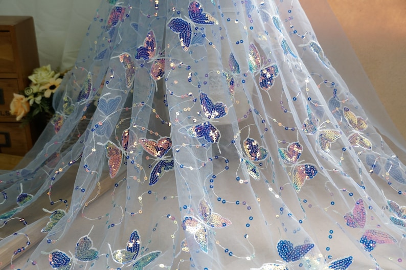 May include: White sheer fabric with iridescent butterfly sequins. The fabric has a delicate, airy feel and is perfect for adding a touch of sparkle to any project.